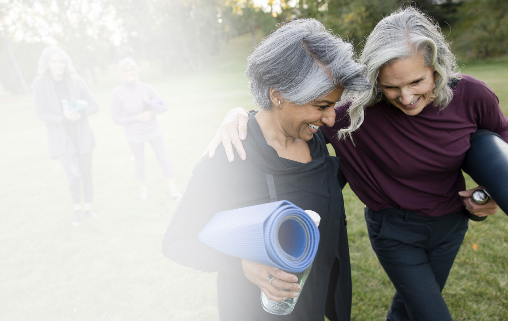 SENIOR WOMEN SMILING ARM IN ARM CARRYING YOGA MATS AT THE PARK